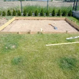 Przygotowanie podłoża pod wylewkę betonową: drewniany szalunek, piasek, łopata i poziomica na trawie, w tle dom jednorodzinny i tuje.