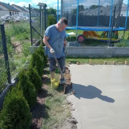 Mężczyzna w niebieskiej koszulce wyrównuje świeżo wylaną betonową wylewkę szpachlą na zewnątrz, obok rosną tuje, a w tle widoczny trampolina i domy w budowie.