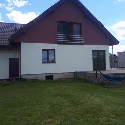 FALBUD - Dwukondygnacyjny dom z jasną elewacją do połowy wysokości i bordową elewacją w górnej części, z widocznym balkonem z metalową barierką i zielonym trawnikiem przed domem.