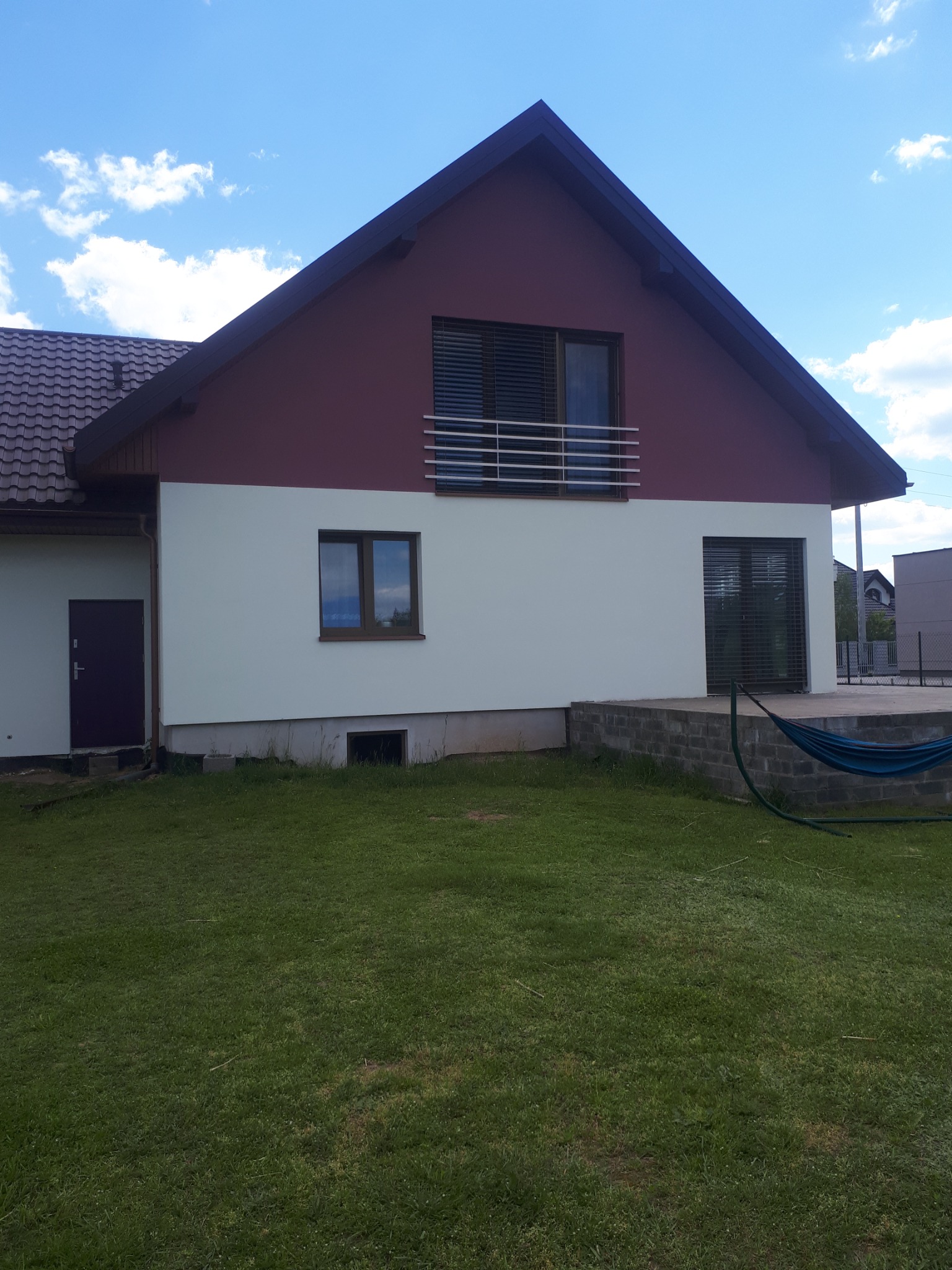 Dwukondygnacyjny dom z jasną elewacją do połowy wysokości i bordową elewacją w górnej części, z widocznym balkonem z metalową barierką i zielonym trawnikiem przed domem.