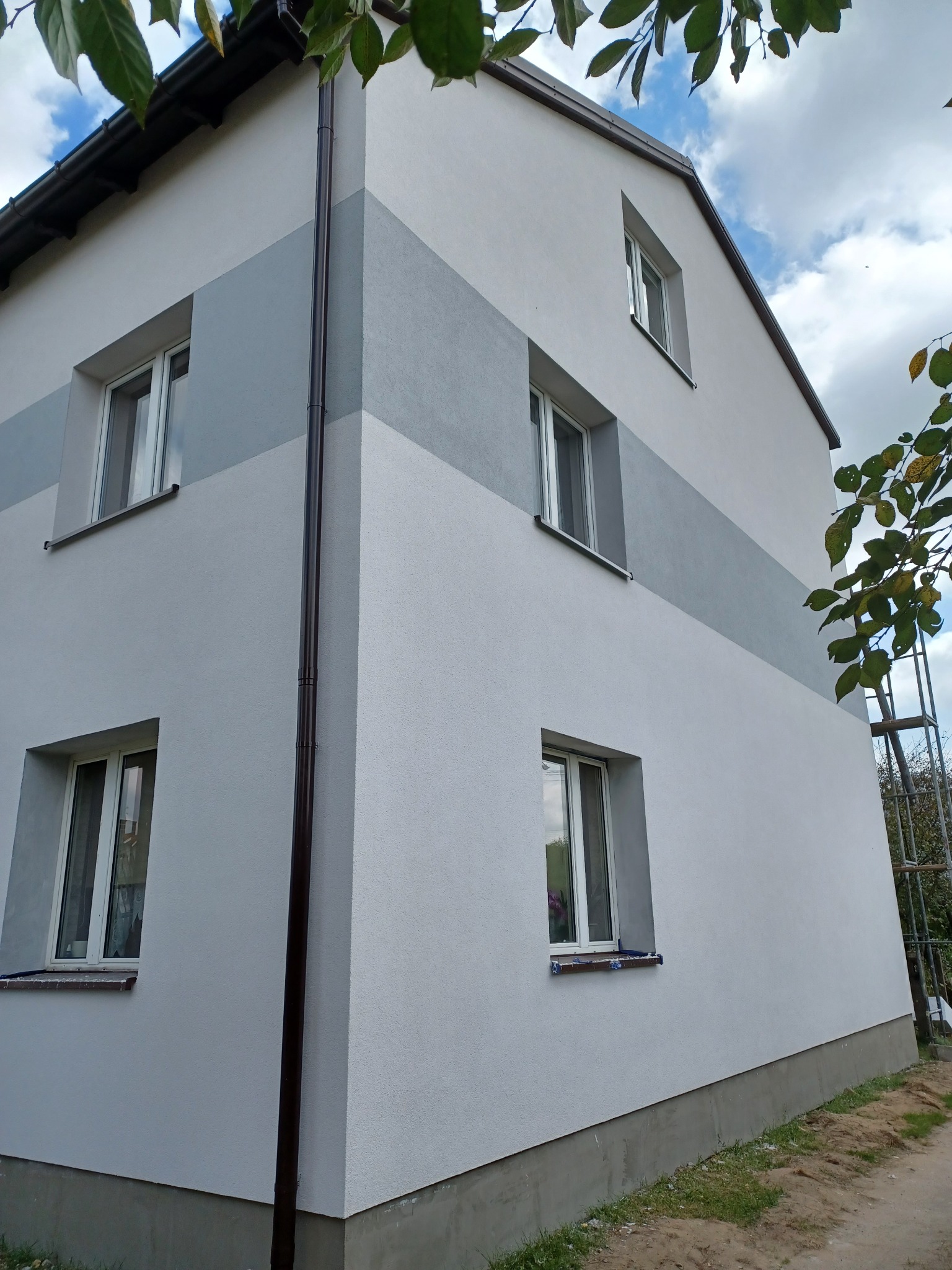 Dwukondygnacyjny dom z oknami po remoncie elewacji, z szarym pasem na środku, widoczny z zewnątrz, z brązową rynną po lewej stronie i rusztowaniem w tle po prawej.