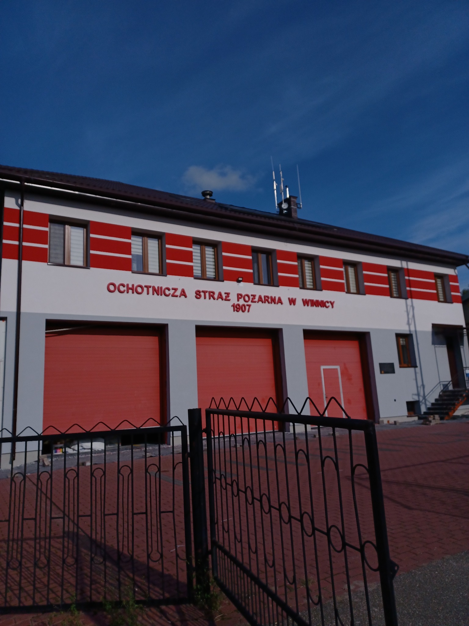 Budynek Ochotniczej Straży Pożarnej w Winnicy po renowacji elewacji, z czerwono-białymi pasami i trzema czerwonymi bramami garażowymi, widziany zza czarnego, metalowego ogrodzenia.