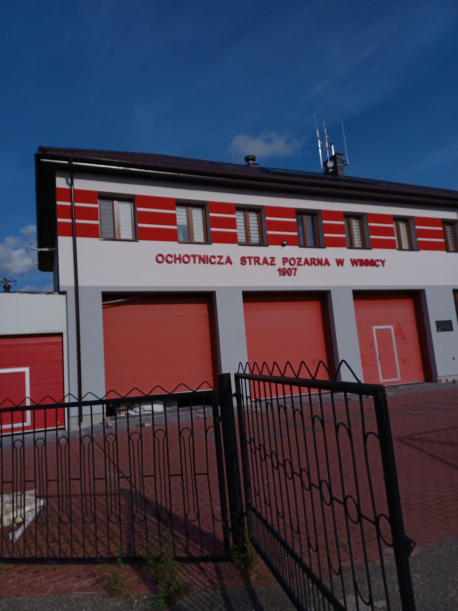 Budynek Ochotniczej Straży Pożarnej w Winnicy z czerwonymi bramami garażowymi i poziomymi pasami na elewacji, widziany zza metalowego ogrodzenia.