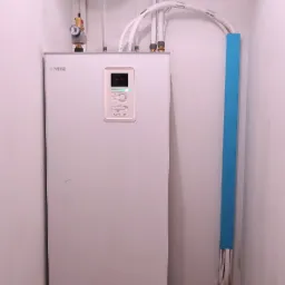 Biała pompa ciepła NIBE BA-SVM 10-200 z niebieskim zbiornikiem wyrównawczym i rurami w narożniku pomieszczenia z brązową podłogą.