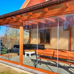 Zadaszony taras z drewnianą konstrukcją i szklanymi ścianami przesuwnymi, wyposażony w meble wypoczynkowe, z widokiem na ogród w słoneczny dzień.