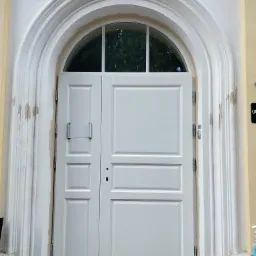 Białe, dwuskrzydłowe drzwi wejściowe z półokrągłym oknem nad nimi, osadzone w łukowatym portalu z dekoracyjnymi pilastrami. Widoczne ślady po montażu na portalu.