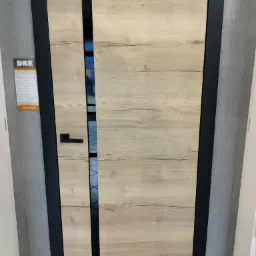 Drzwi wewnętrzne DRE z okleiną imitującą jasne drewno, z czarnymi elementami ramy i klamką, widoczne przez szparę wnętrze.