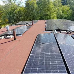 Instalacja paneli fotowoltaicznych na dachu pokrytym czerwoną papą, widok z góry, kominki wentylacyjne w tle drzewa.