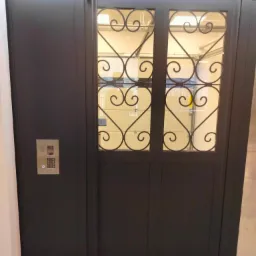 Ciemna brama garażowa z ozdobnymi, metalowymi elementami w górnej części i panelem z kodem dostępu po lewej stronie.