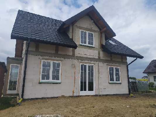 Dom w trakcie budowy z zamontowanymi białymi oknami, widoczne ocieplenie i czarna dachówka.