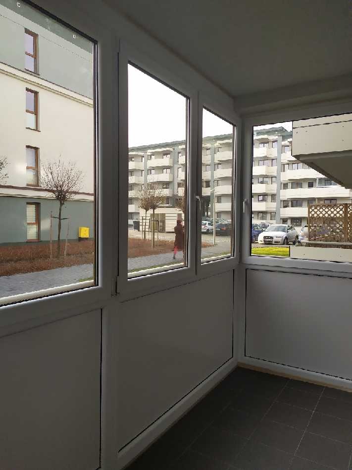 Zabudowa balkonu z białymi ramami okiennymi i widokiem na osiedle mieszkaniowe z chodnikiem i przechodzącą osobą.