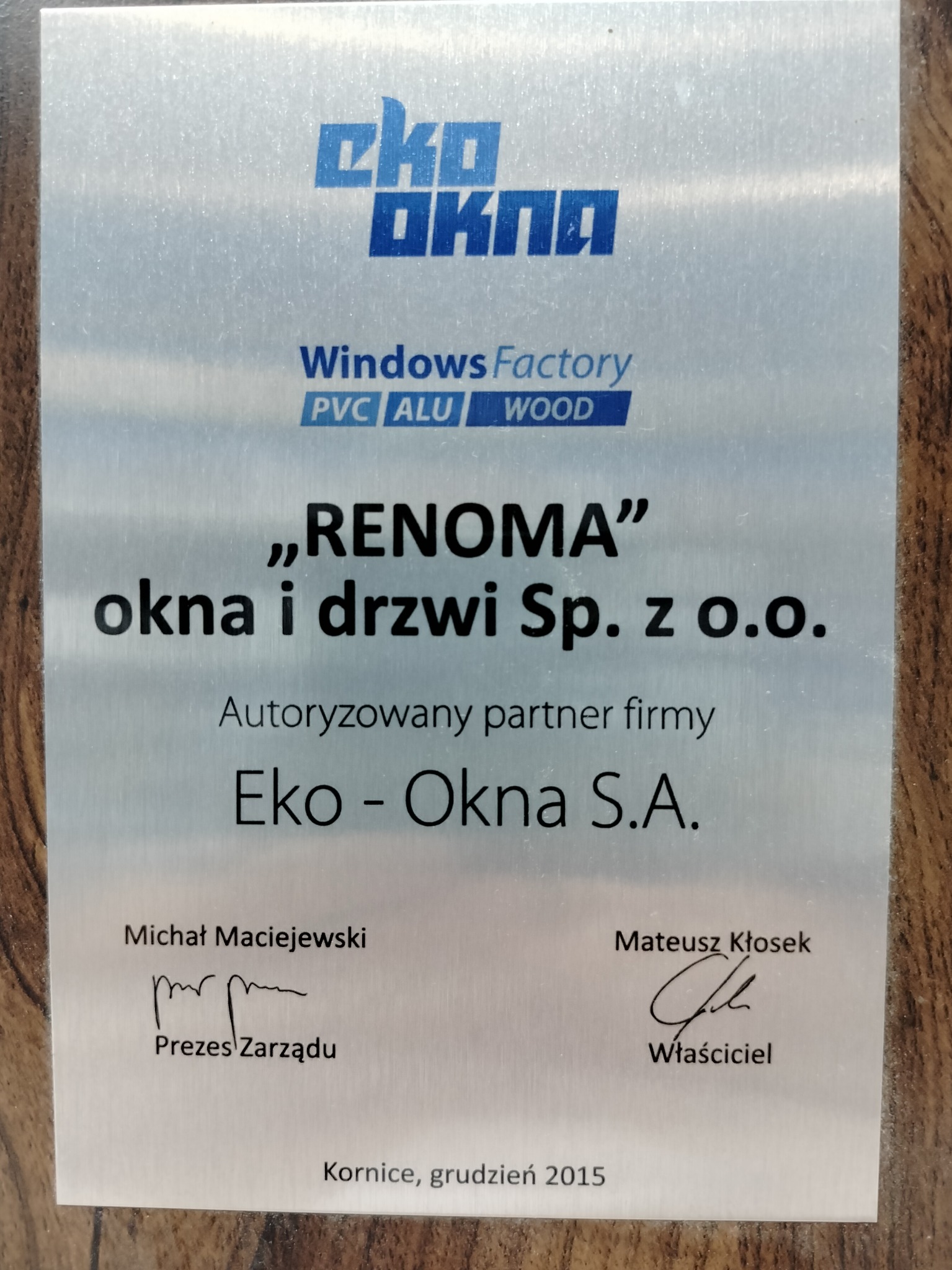 Metalowa tabliczka informacyjna firmy Renoma, autoryzowanego partnera Eko-Okna S.A., z logo Eko-Okna, adresem firmy i podpisami Prezesa Zarządu i Właściciela, umieszczona na drewnianym tle.