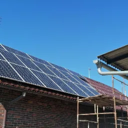 Częściowy widok domu z cegły z panelami słonecznymi na dachu i rusztowaniem po prawej stronie, błękitne niebo w tle.