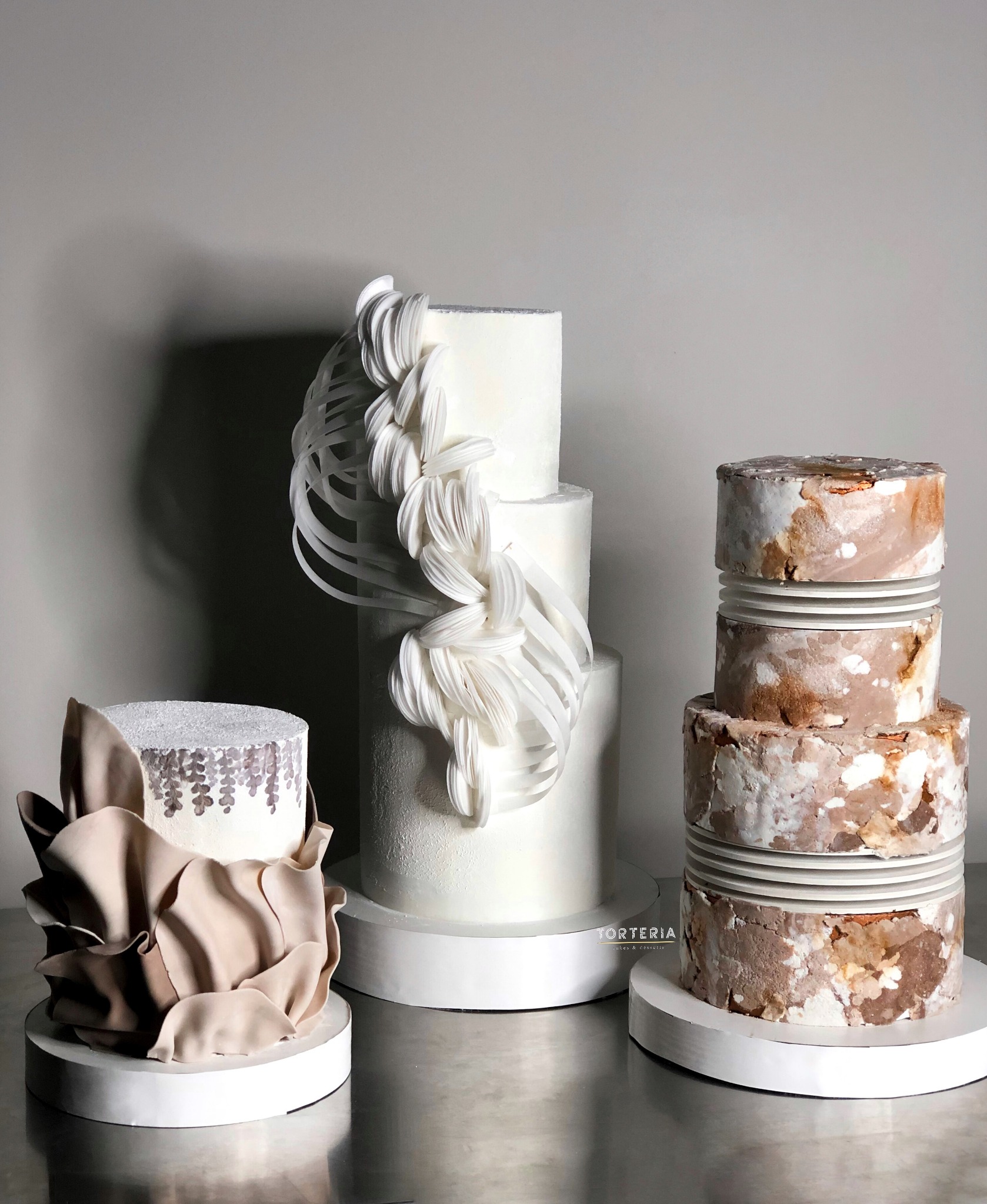 Trzy torty artystyczne: jeden z beżowymi falbanami, drugi biały z abstrakcyjną dekoracją z białych wstążek, trzeci w stylu rustykalnym z efektem marmuru w odcieniach brązu i bieli.