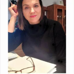 Kobieta w czarnym swetrze siedzi za biurkiem z dokumentami i okularami, trzymając długopis w uniesionej dłoni, w tle logo MetLife i drewniana szafka.