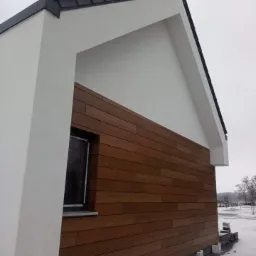 Fragment nowo wybudowanego domu z elewacją z paneli drewnopodobnych, biały tynk powyżej, widoczne ośnieżone otoczenie.