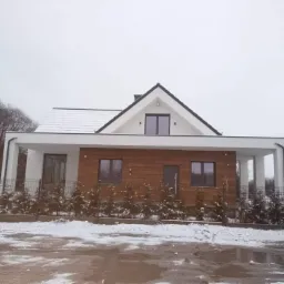 Nowoczesny dom jednorodzinny z płaskim dachem pokrytym śniegiem, elewacja z elementami drewnopodobnymi i białymi kolumnami podtrzymującymi zadaszenie nad wejściem, zimowa sceneria.