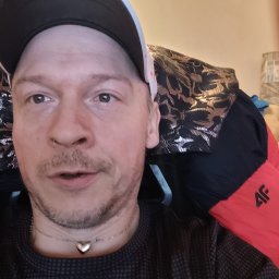 PRZEDSIĘBIORSTWO AUTOBUSOWE KONCESJONOWANE GNIEZNO - STANISŁAWÓW - Portret mężczyzny w czapce z daszkiem, ubranego w ciemną koszulę w kropki i złoty naszyjnik z sercem, siedzącego na krześle z wzorzystym obiciem i czerwoną kurtką 4F.