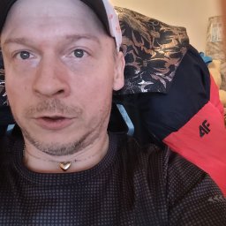 PRZEDSIĘBIORSTWO AUTOBUSOWE KONCESJONOWANE GNIEZNO - STANISŁAWÓW - Autoportret mężczyzny w czapce z daszkiem i naszyjnikiem w kształcie serca, siedzącego na krześle z wzorzystym oparciem i czerwonym zagłówkiem.