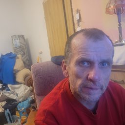 PRZEDSIĘBIORSTWO AUTOBUSOWE KONCESJONOWANE GNIEZNO - STANISŁAWÓW - Autoportret mężczyzny w czerwonej bluzie na tle pokoju z widocznym bałaganem i elementami wyposażenia.