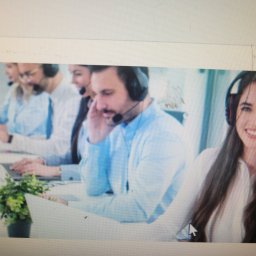 PRZEDSIĘBIORSTWO AUTOBUSOWE KONCESJONOWANE GNIEZNO - STANISŁAWÓW - Zespół konsultantów w słuchawkach pracuje przy biurkach. Kobieta z uśmiechem patrzy w kamerę. Widoczny fragment rośliny doniczkowej. Jasne, nowoczesne biuro.