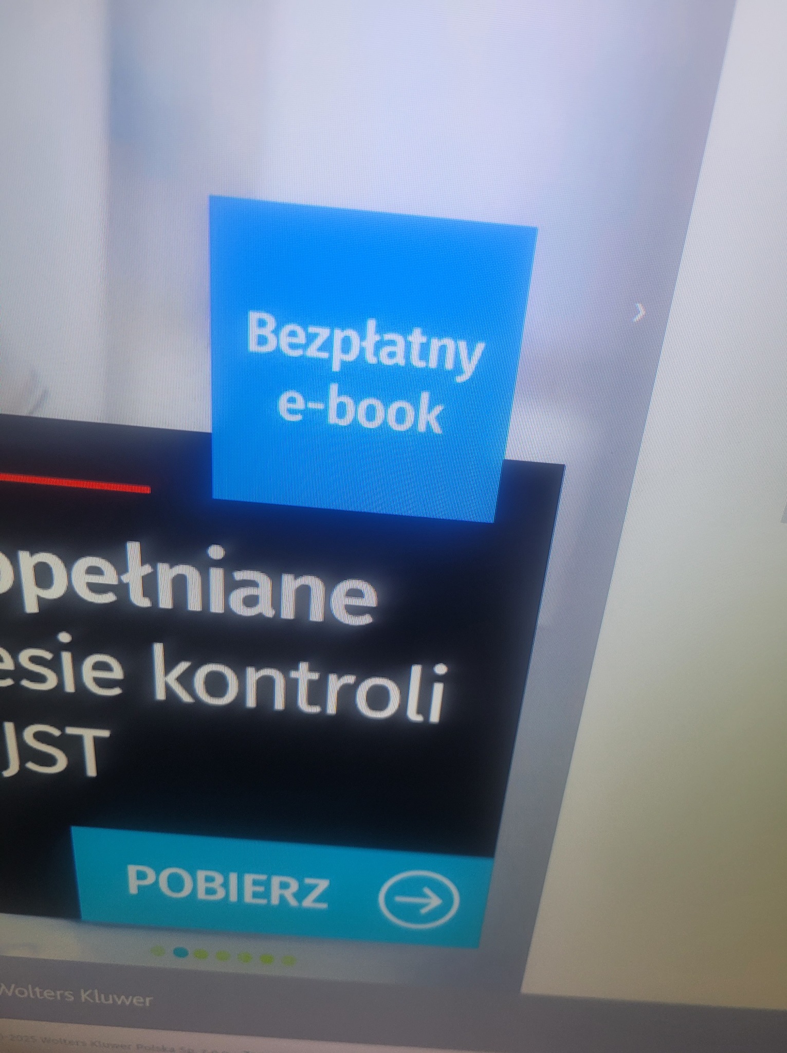 Ekran monitora z ofertą darmowego e-booka o kontroli JST, z przyciskiem 'Pobierz' i logo Wolters Kluwer w dolnej części. Ujęcie z bliska, lekko zniekształcone.