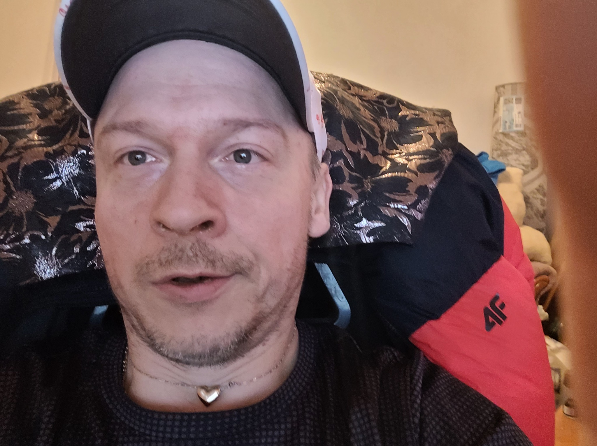 Portret mężczyzny w czapce z daszkiem, ubranego w ciemną koszulę w kropki i złoty naszyjnik z sercem, siedzącego na krześle z wzorzystym obiciem i czerwoną kurtką 4F.