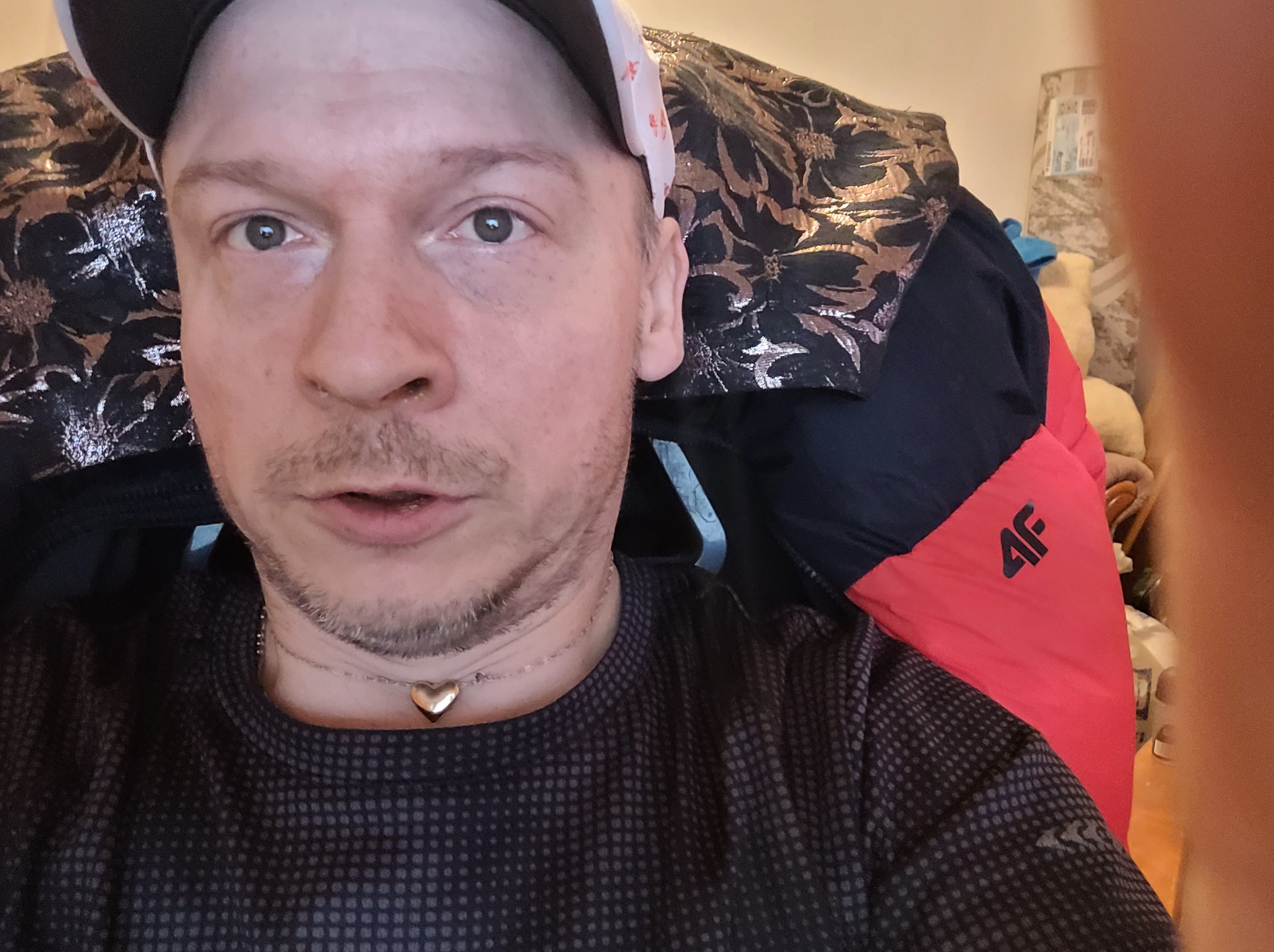 Autoportret mężczyzny w czapce z daszkiem i naszyjnikiem w kształcie serca, siedzącego na krześle z wzorzystym oparciem i czerwonym zagłówkiem.