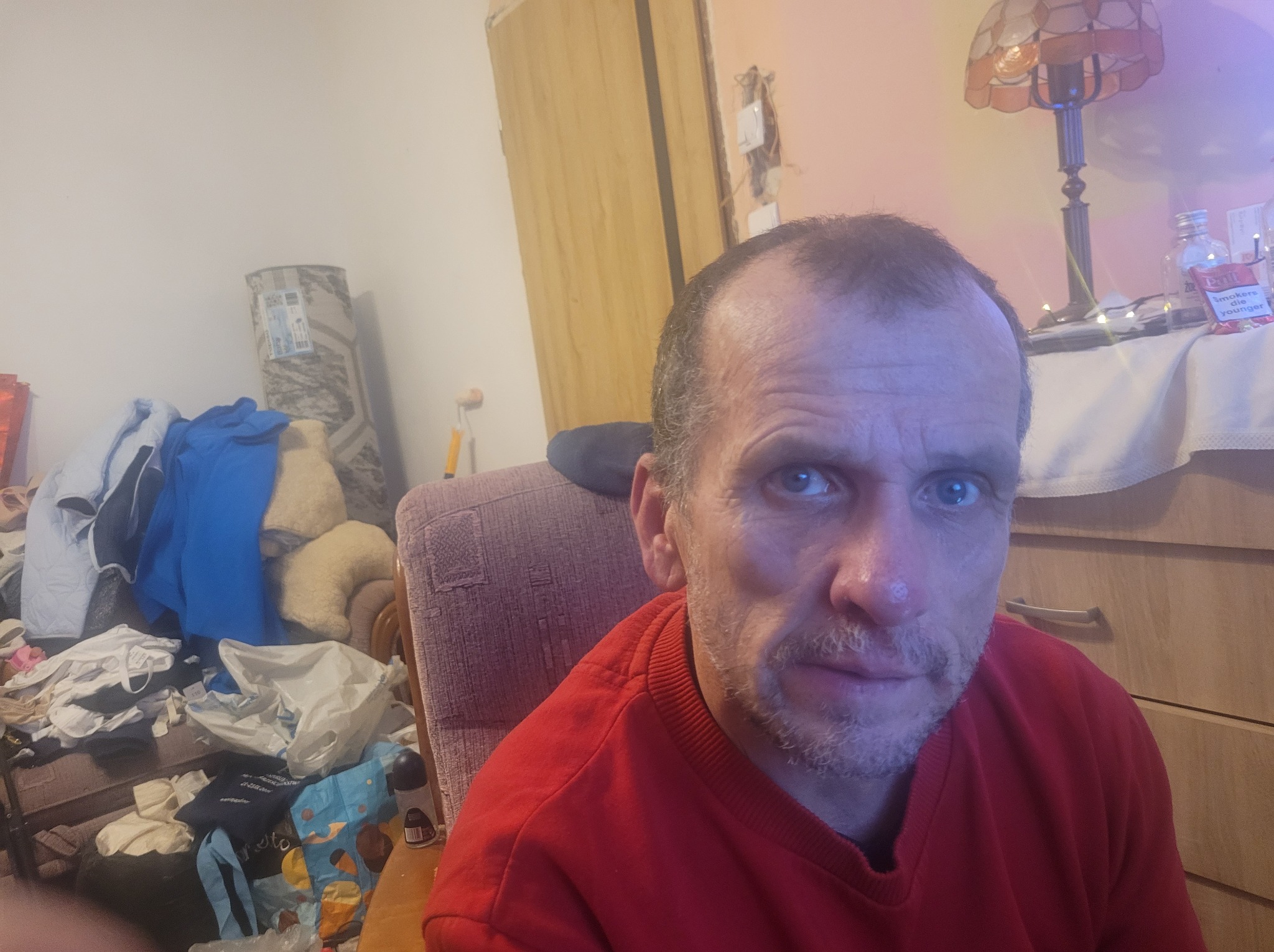 Autoportret mężczyzny w czerwonej bluzie na tle pokoju z widocznym bałaganem i elementami wyposażenia.