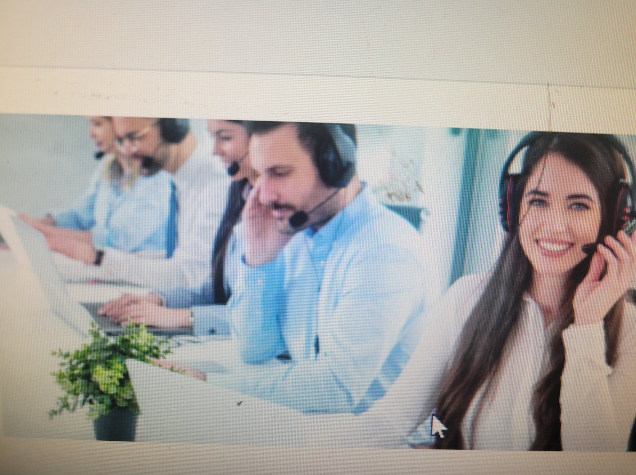 Zespół konsultantów w słuchawkach pracuje przy biurkach. Kobieta z uśmiechem patrzy w kamerę. Widoczny fragment rośliny doniczkowej. Jasne, nowoczesne biuro.