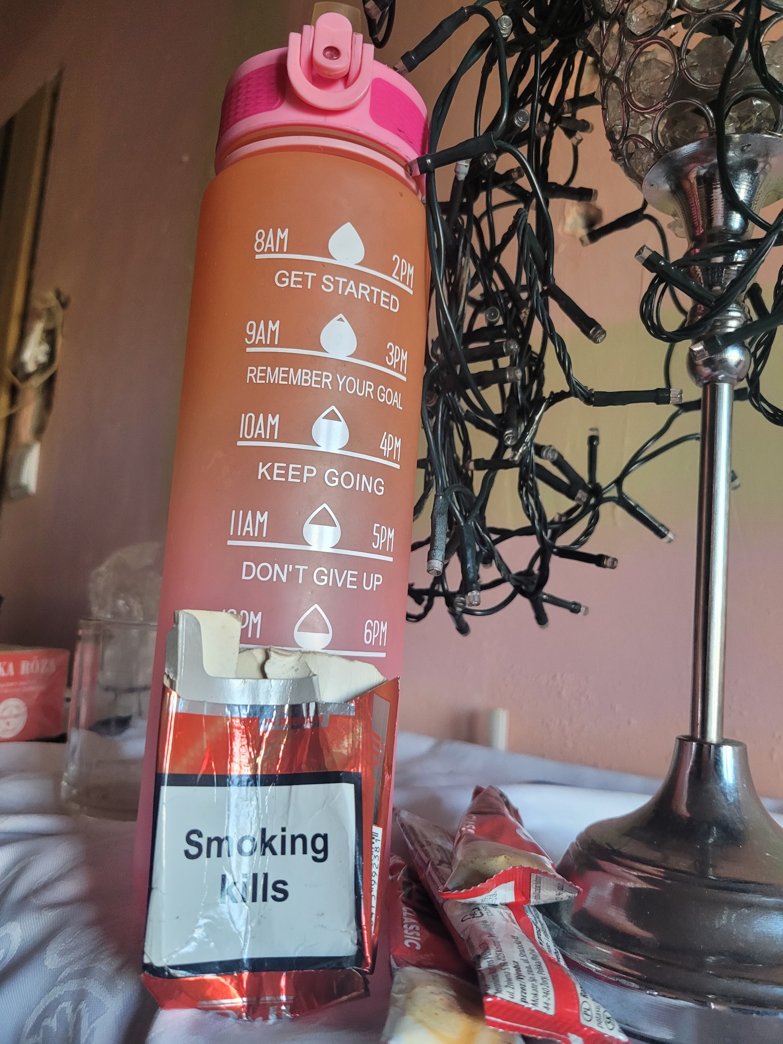 Nietypowe ujęcie: bidon z motywacyjnymi napisami, paczka papierosów z napisem 'Smoking Kills', lampka nocna i girlanda świetlna na stole.