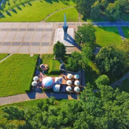 Widok z lotu ptaka na park z pomnikiem, placem i ogródkiem piwnym z parasolami, otoczony zielenią.