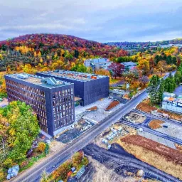 Zdjęcie z drona przedstawia dwa nowoczesne budynki biurowe z ciemną elewacją, otoczone jesiennym krajobrazem z kolorowymi drzewami i placem budowy w trakcie prac wykończeniowych.