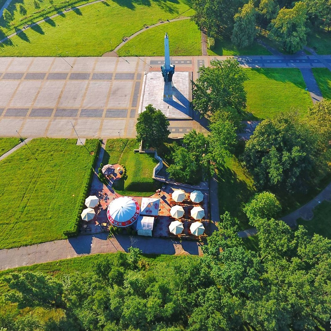 Widok z lotu ptaka na park z pomnikiem, placem i ogródkiem piwnym z parasolami, otoczony zielenią.