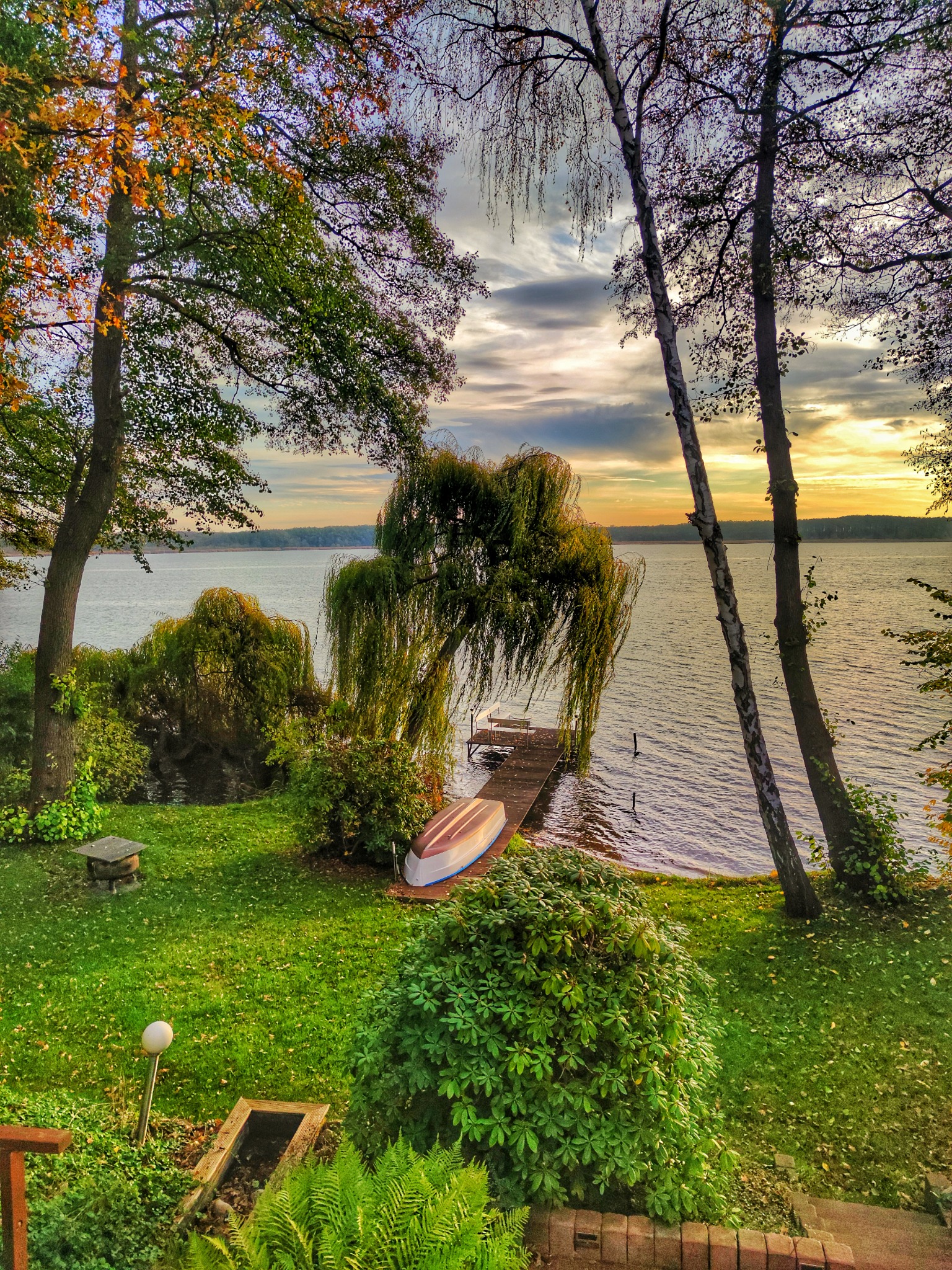 Jesienny krajobraz jeziora w Tychach o zachodzie słońca, z łodzią na brzegu i pomostem, widok z ogrodu.