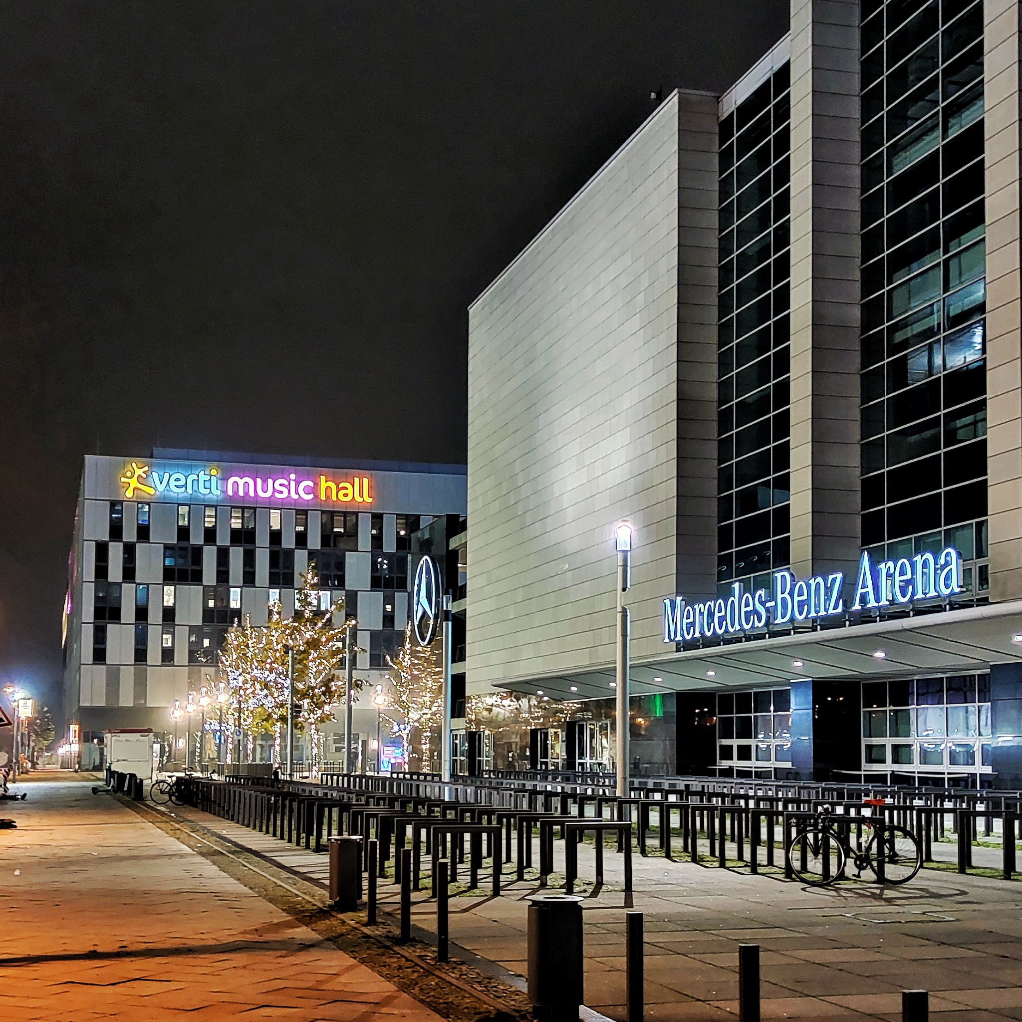 Nocny widok na Mercedes-Benz Arena i Verti Music Hall z oświetlonymi drzewami, widoczne stojaki na rowery i chodnik z kostki.