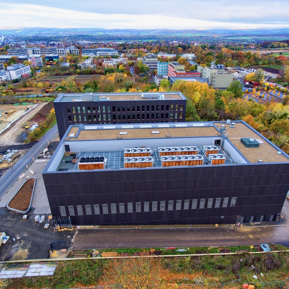 Widok z góry na nowoczesny budynek z licznymi klimatyzatorami na dachu, w tle zabudowa miejska i jesienne drzewa.