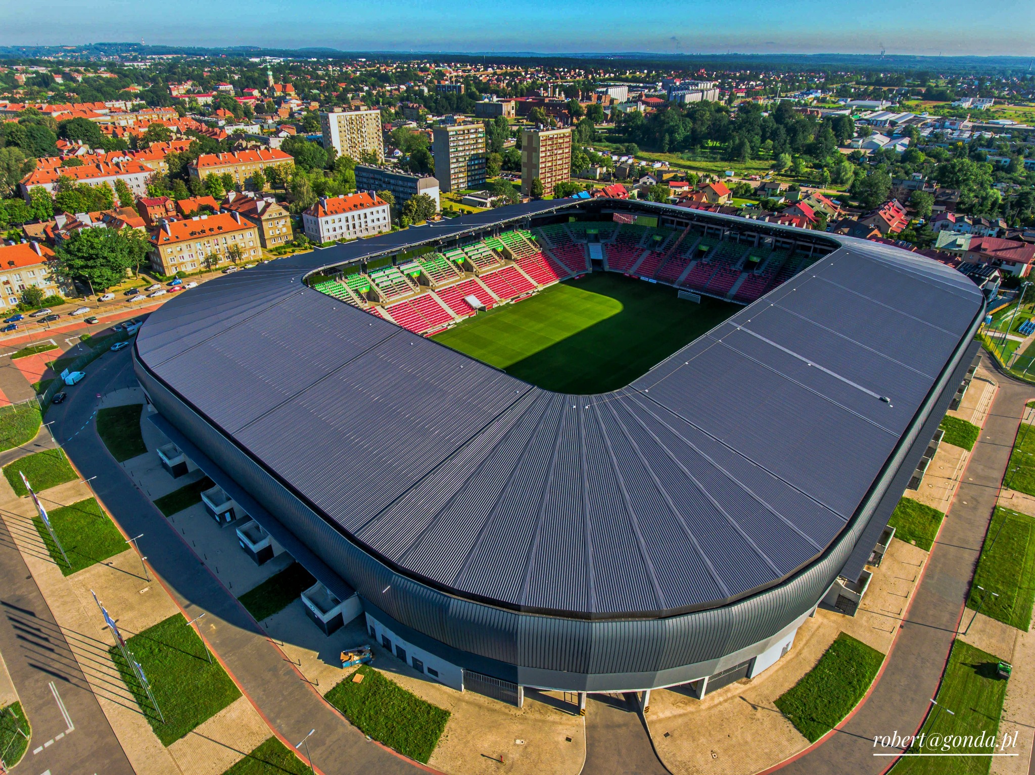 Panoramiczny widok z lotu ptaka na nowoczesny stadion piłkarski z zieloną murawą i czerwono-zielonymi trybunami, otoczony miejską zabudową, w słoneczny dzień.