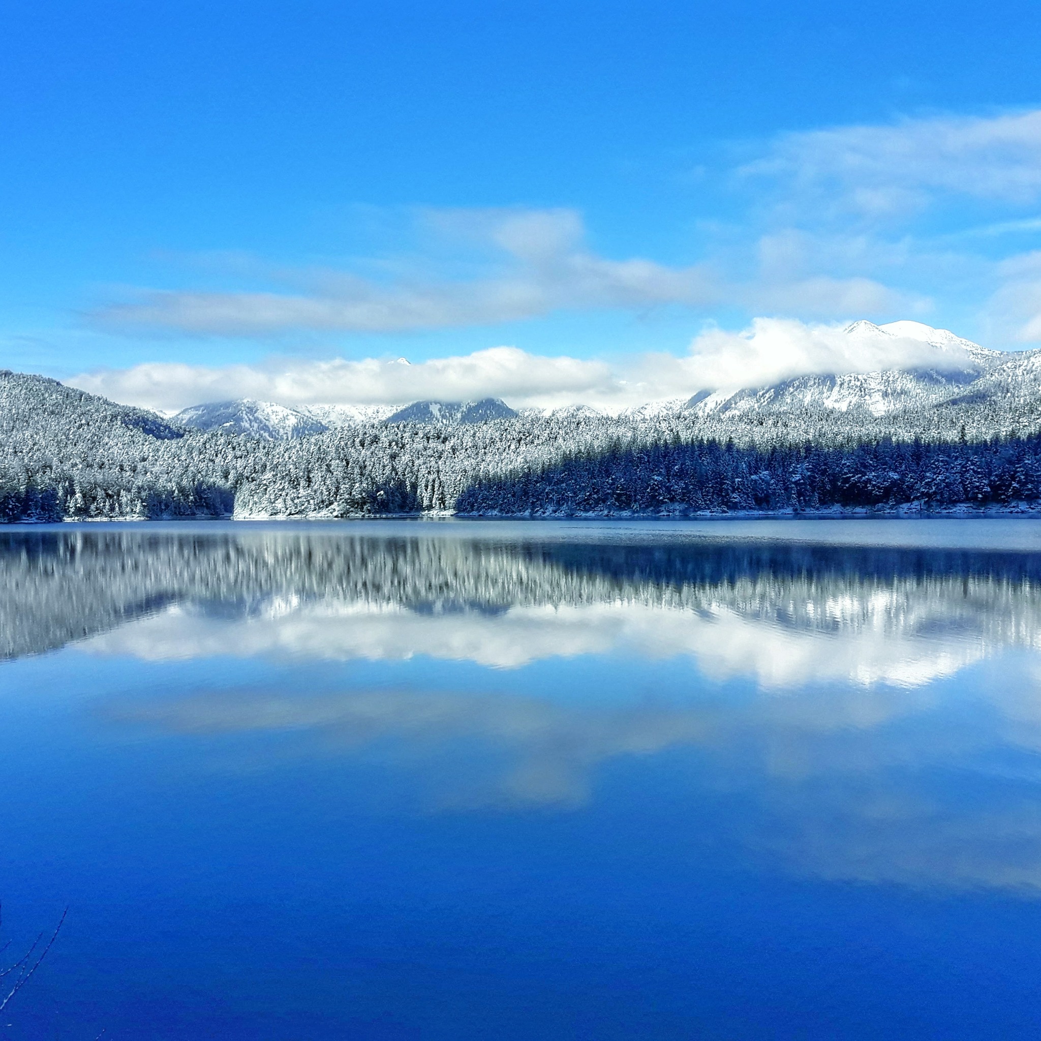 Zimowy krajobraz: błękitne jezioro odbija zaśnieżone góry i las pod jasnym, błękitnym niebem z delikatnymi obłokami.