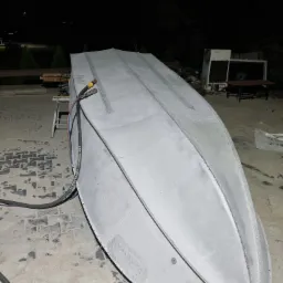 Odwrócona aluminiowa łódź po piaskowaniu, widoczne ślady po procesie na powierzchni i betonowym podłożu, w tle zieleń i elementy wyposażenia warsztatowego, przewód do piaskowania leżący obok.