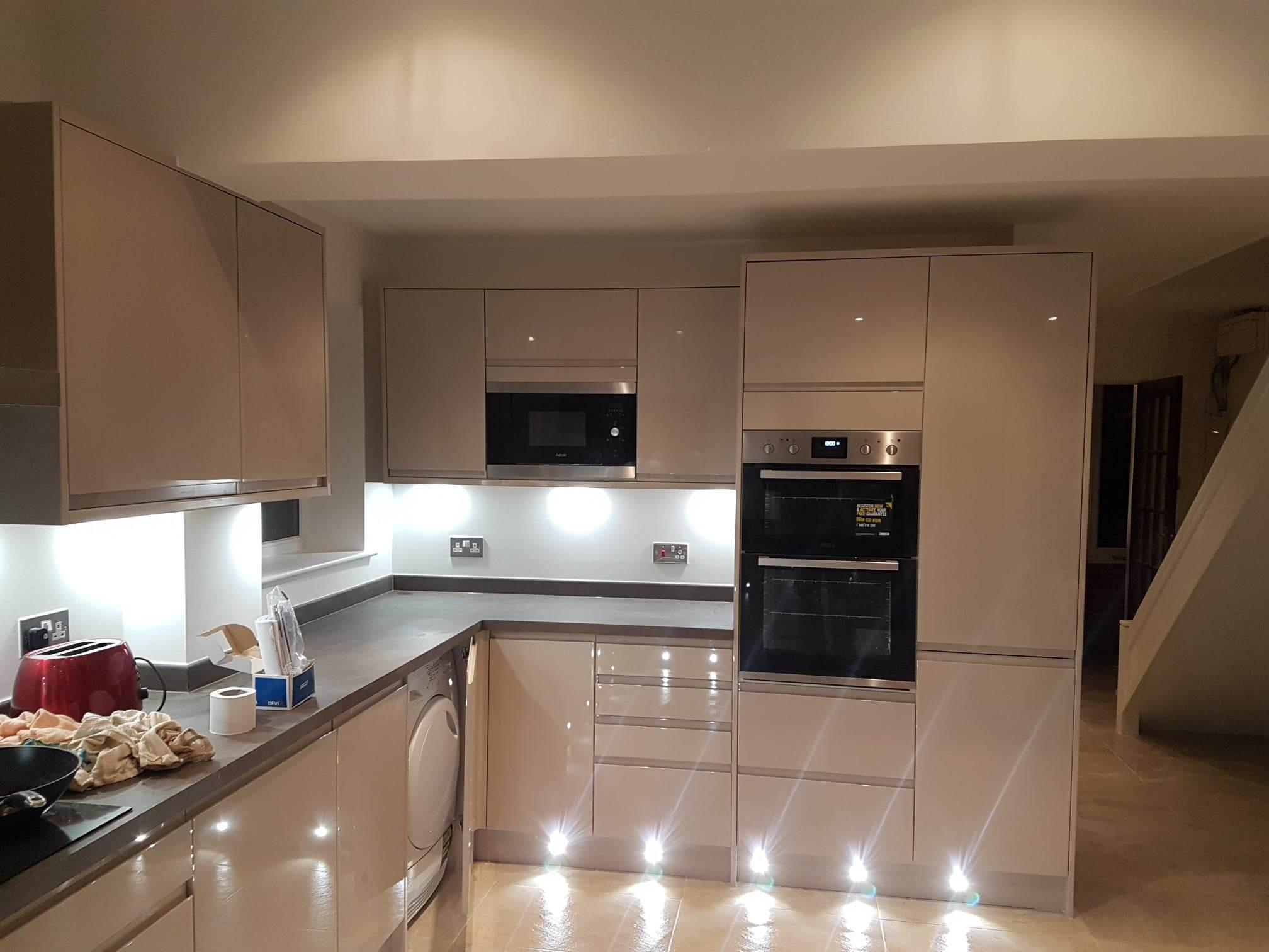 Nowoczesna kuchnia z beżowymi, lakierowanymi frontami, oświetleniem podszafkowym i podłogowym, widoczny sprzęt AGD: piekarnik, mikrofalówka i pralka.