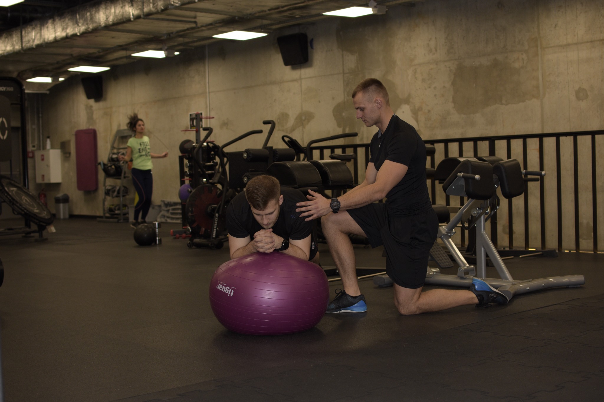 Mężczyzna ćwiczy na fioletowej piłce Tiguan w siłowni, a drugi mężczyzna w czarnym stroju go asekuruje. W tle kobieta skacze na skakance.