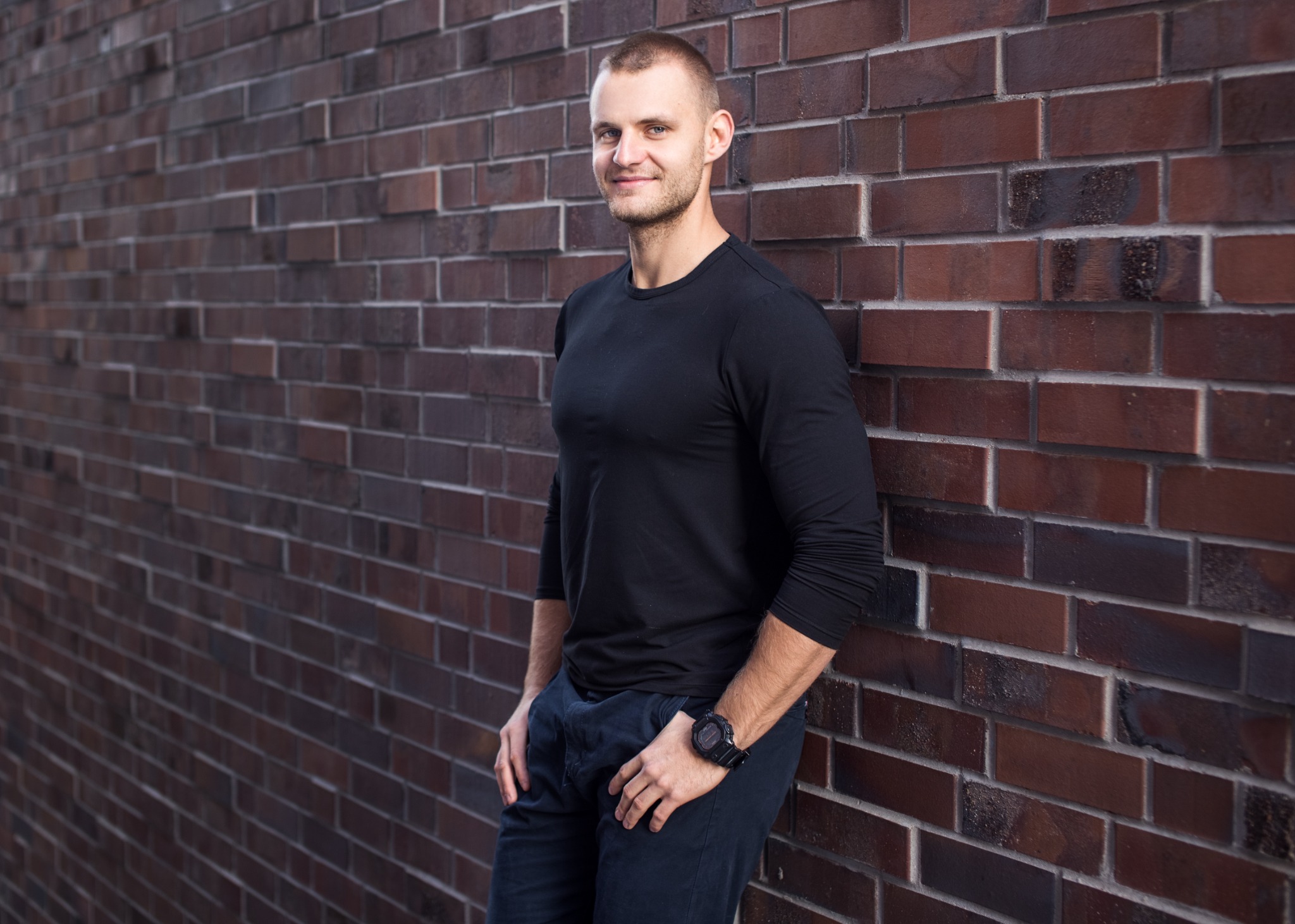 Mężczyzna w czarnej koszulce z długim rękawem oparty o ceglaną ścianę, z widocznym zegarkiem na nadgarstku i lekkim uśmiechem.