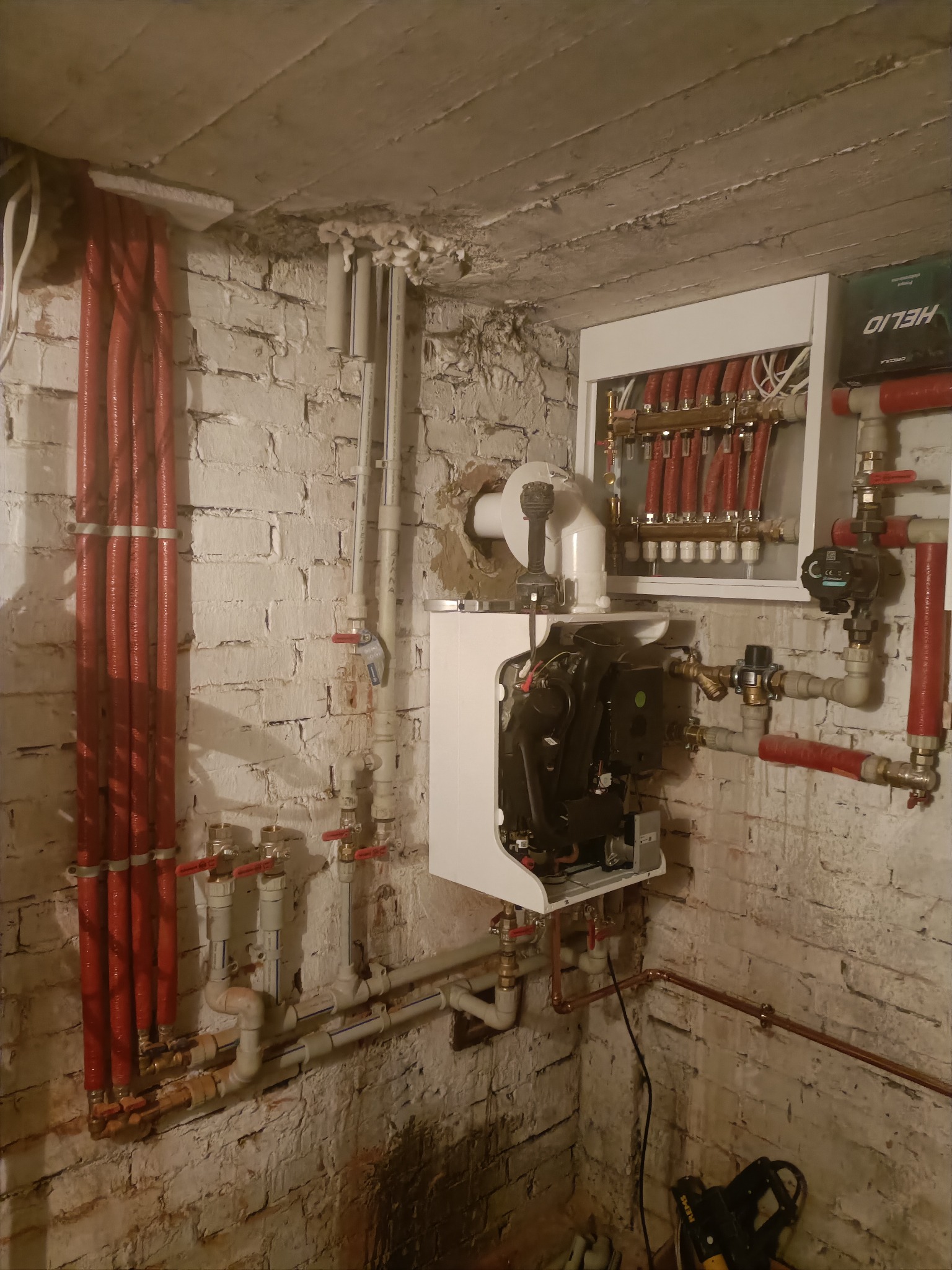 Instalacja kotła grzewczego z rozdzielaczem na tle ceglanej ściany. Widoczne rury w izolacji, zawory i elementy sterujące. Częściowo widoczny sprzęt instalacyjny.