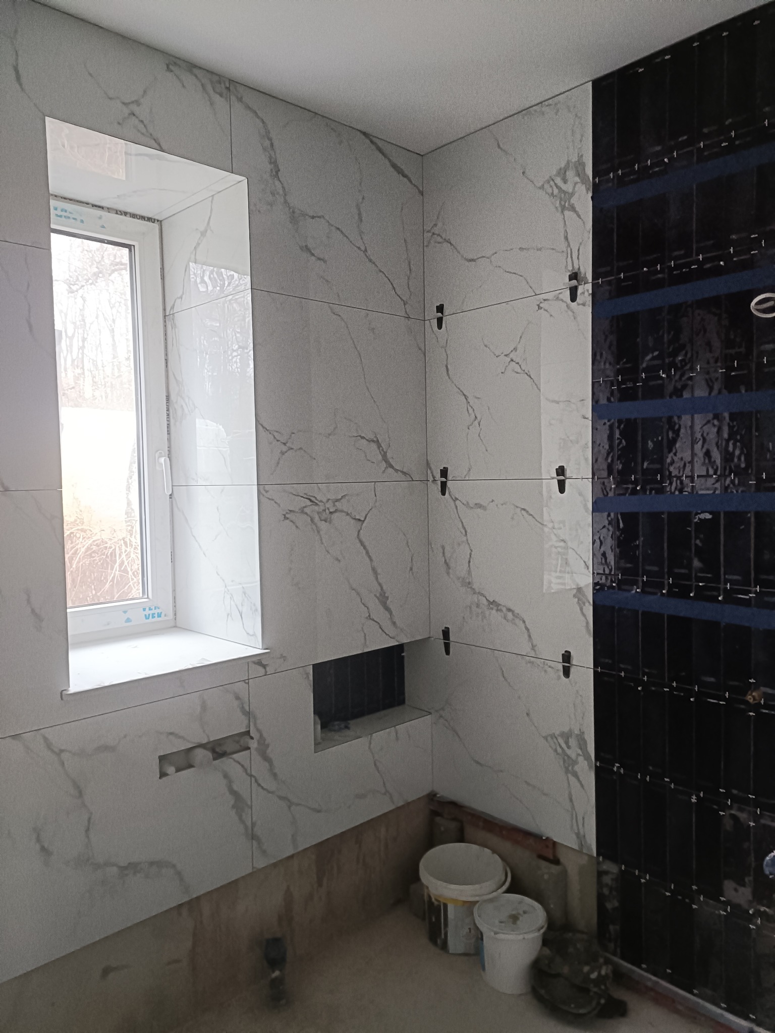 Wnętrze łazienki w trakcie wykańczania: białe płytki imitujące marmur na ścianach wokół okna i czarne, wąskie płytki ułożone pionowo na sąsiedniej ścianie, z widocznymi krzyżykami dystansowymi.