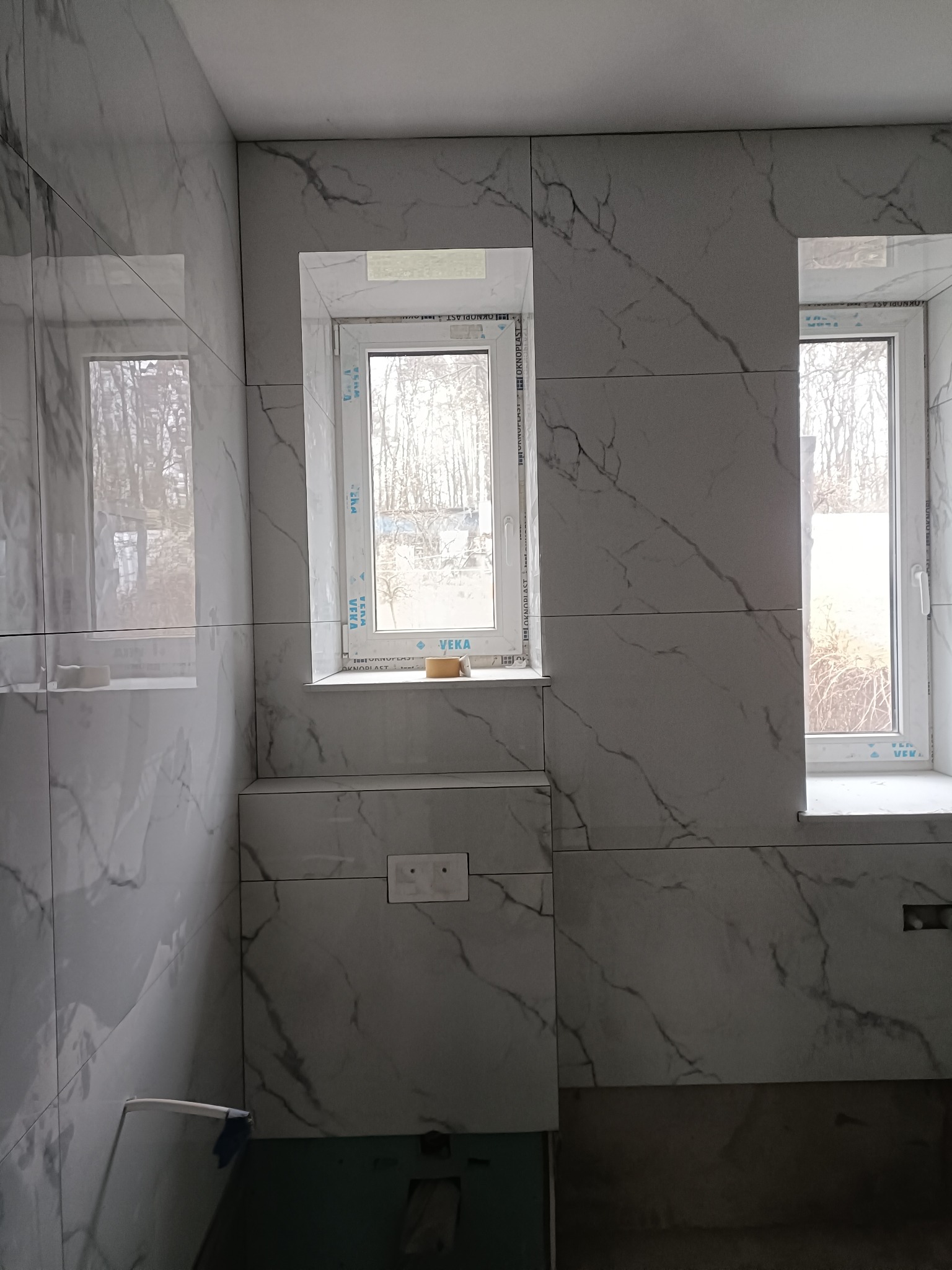 Wykończona łazienka z marmurowymi płytkami, widoczne dwa okna z białymi ramami. Na parapecie leży gąbka. Minimalistyczny styl, jasne wnętrze.