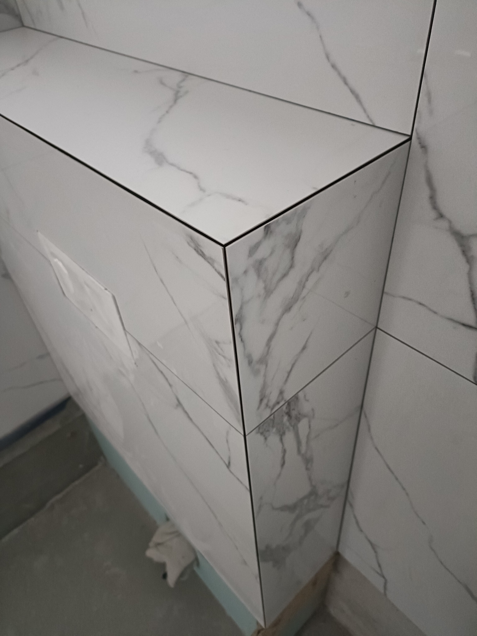Białe płytki imitujące marmur, ułożone pod kątem, tworzące narożnik. Widoczne fugi w kolorze czarnym. Minimalistyczny design, precyzyjne wykończenie.