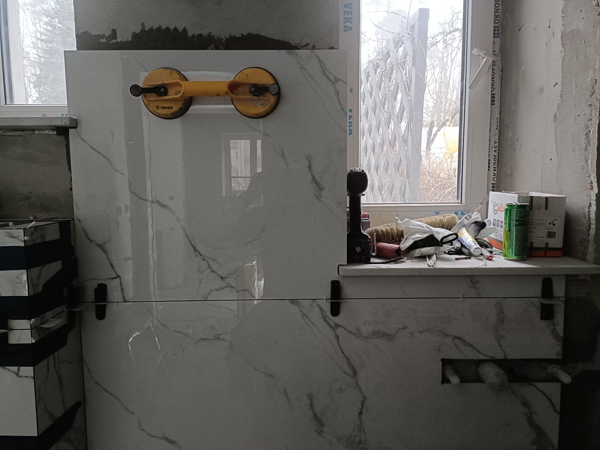 Białe płytki imitujące marmur, układane na ścianie w łazience, z przyssawką do płytek i narzędziami na parapecie okiennym. Widoczne krzyżyki dystansowe.