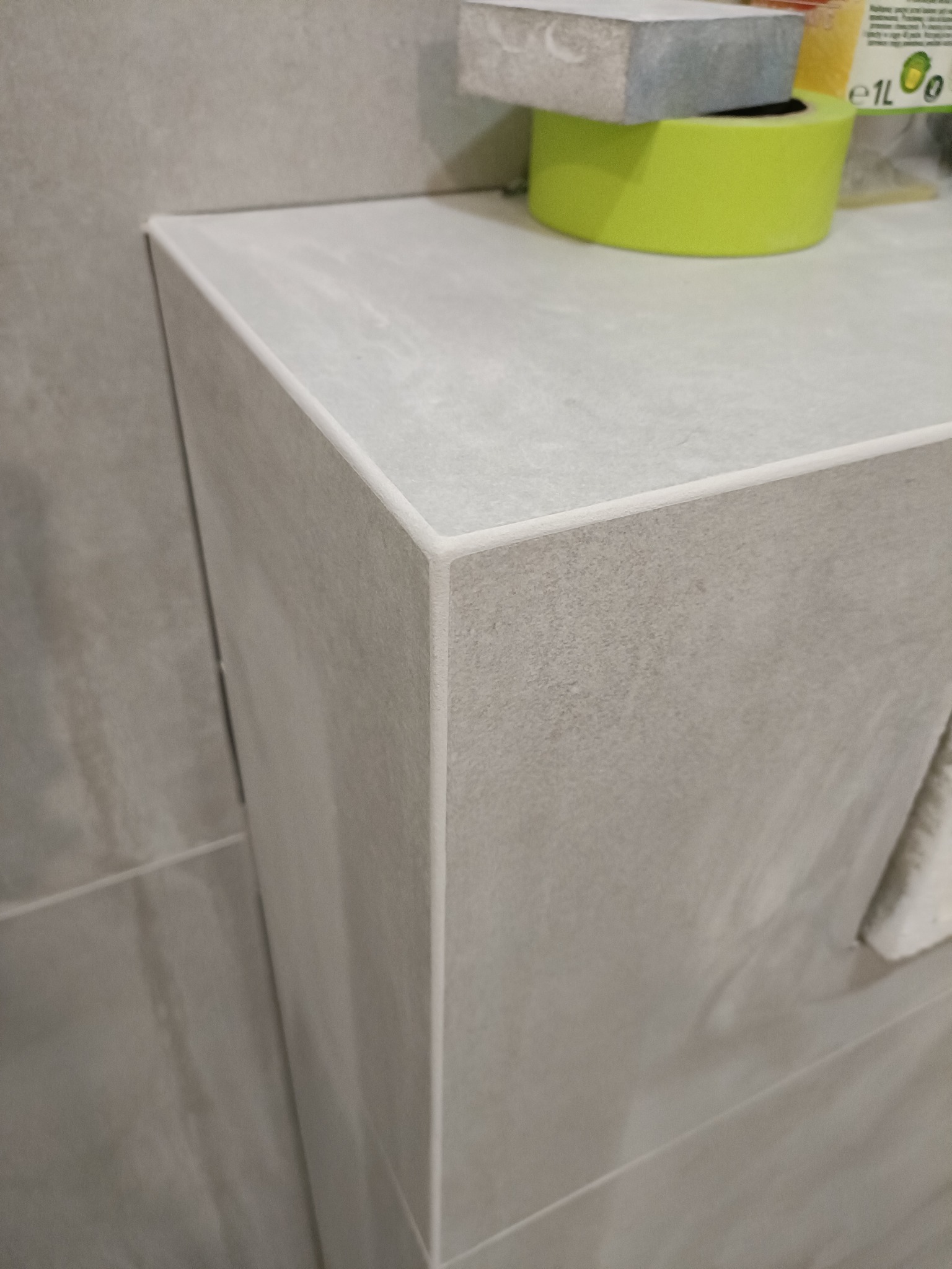 Wykończona półka w łazience z szarych płytek, widoczne detale łączeń i fug. Na górze leżą narzędzia: gąbka i taśma malarska.