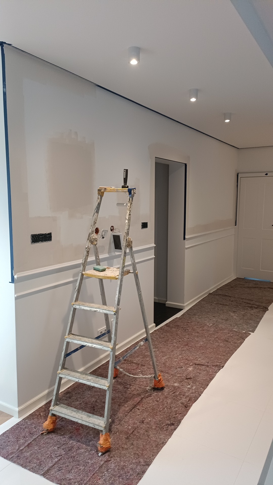 Wnętrze w trakcie malowania: drabina oparta o ścianę z listwami, zabezpieczona folią podłoga, ściana z fragmentami pomalowanymi i taśmą malarską. Widoczne gniazdka elektryczne.